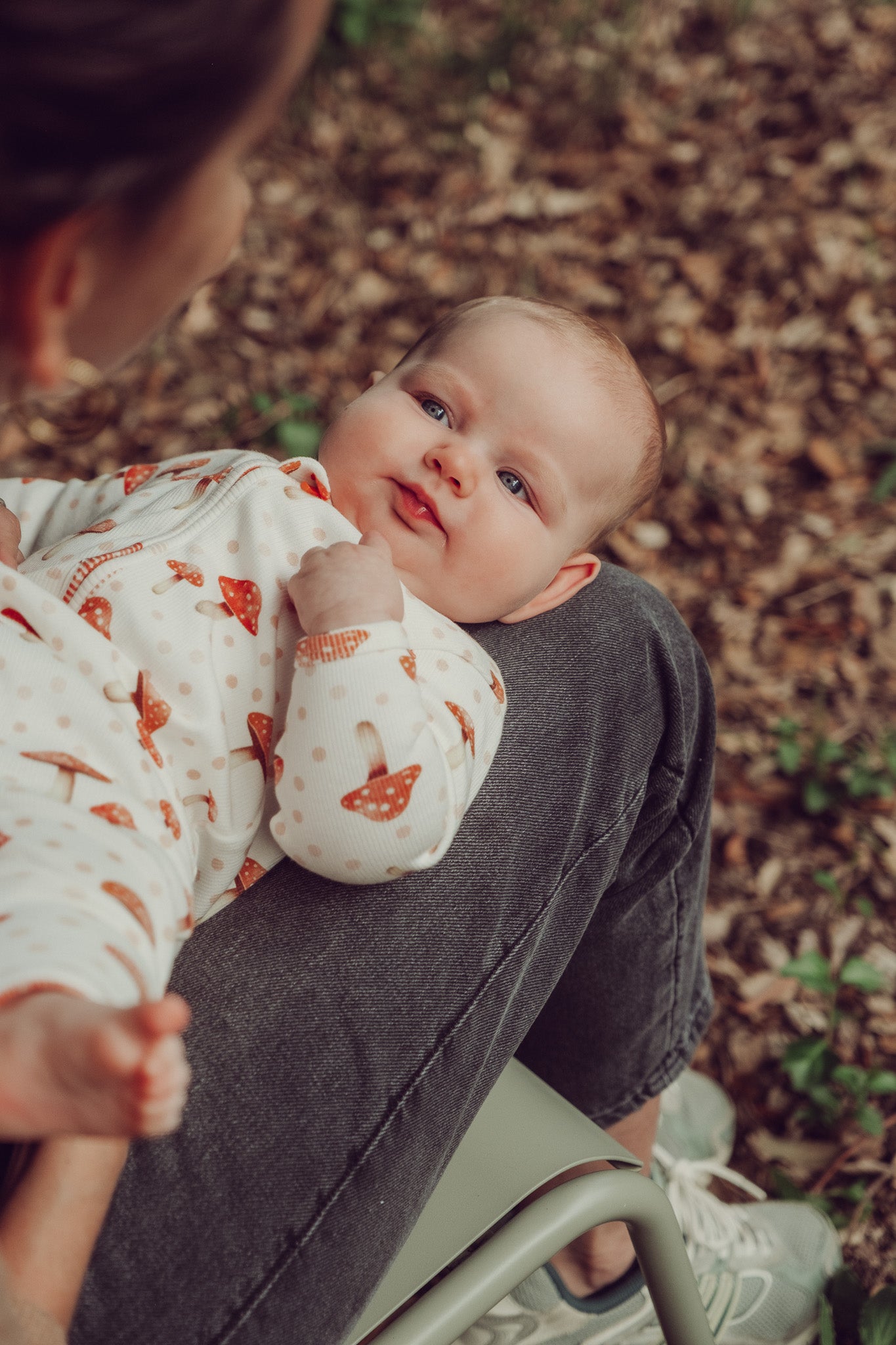 Baby in a floral outfit being held by an adult outdoors on a leafy ground.